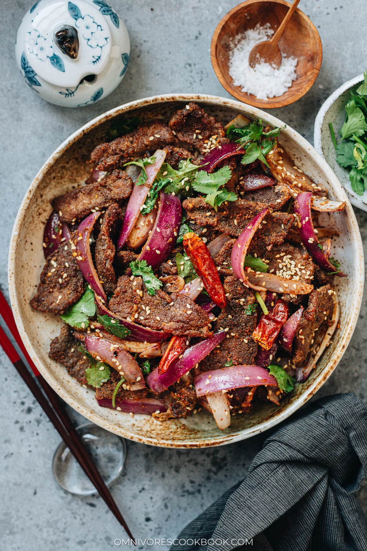 A close-up of cumin beef in a ceramic bowl with red onion, cilantro, and sesame seeds.