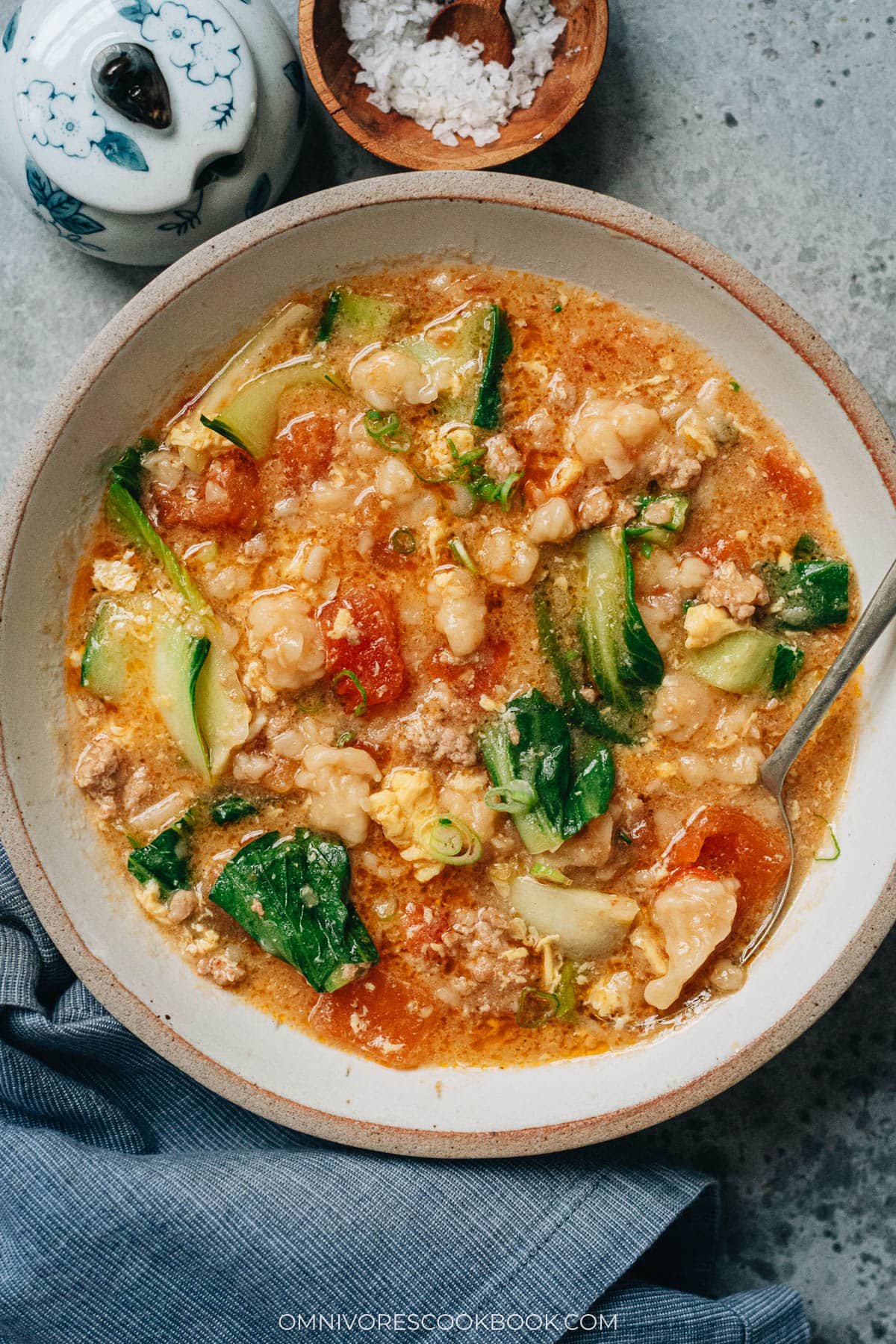 A bowl of Ge Da Tang served on a table, filled with chewy dough drops, wilted bok choy, ground pork, and soft egg ribbons in a tomato-based broth.